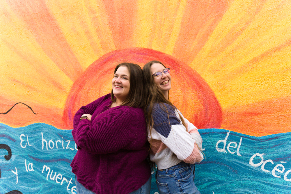 Two people stand back-to-back in front of a colorful mural depicting a bright orange and pink sunset over blue ocean waves. The person on the left, Nicole, wears a deep purple sweater and smiles at the camera with her arms crossed. The person on the right, Emma, wears glasses and a striped blue, white, and pink sweater with jeans, and smiles at the camera.